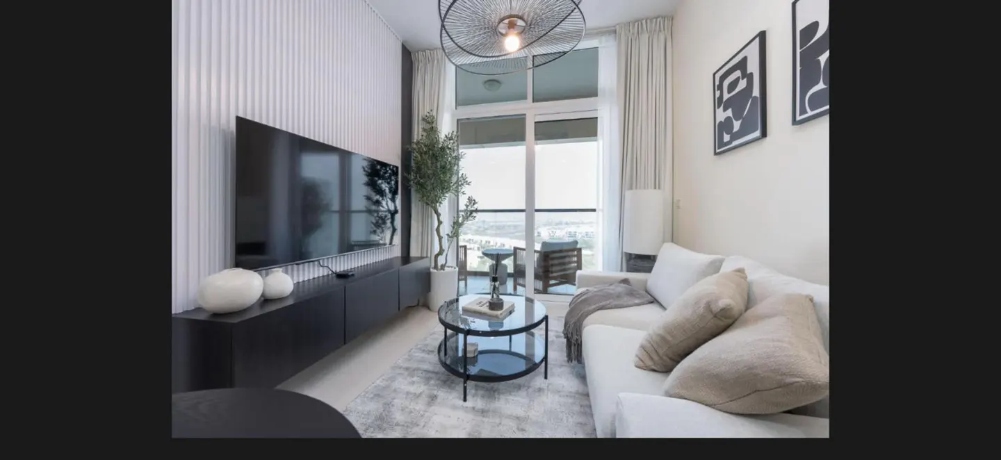 Living room with a white sofa, black TV stand, and a round glass coffee table. Balcony with a chair is visible through the sliding glass door.