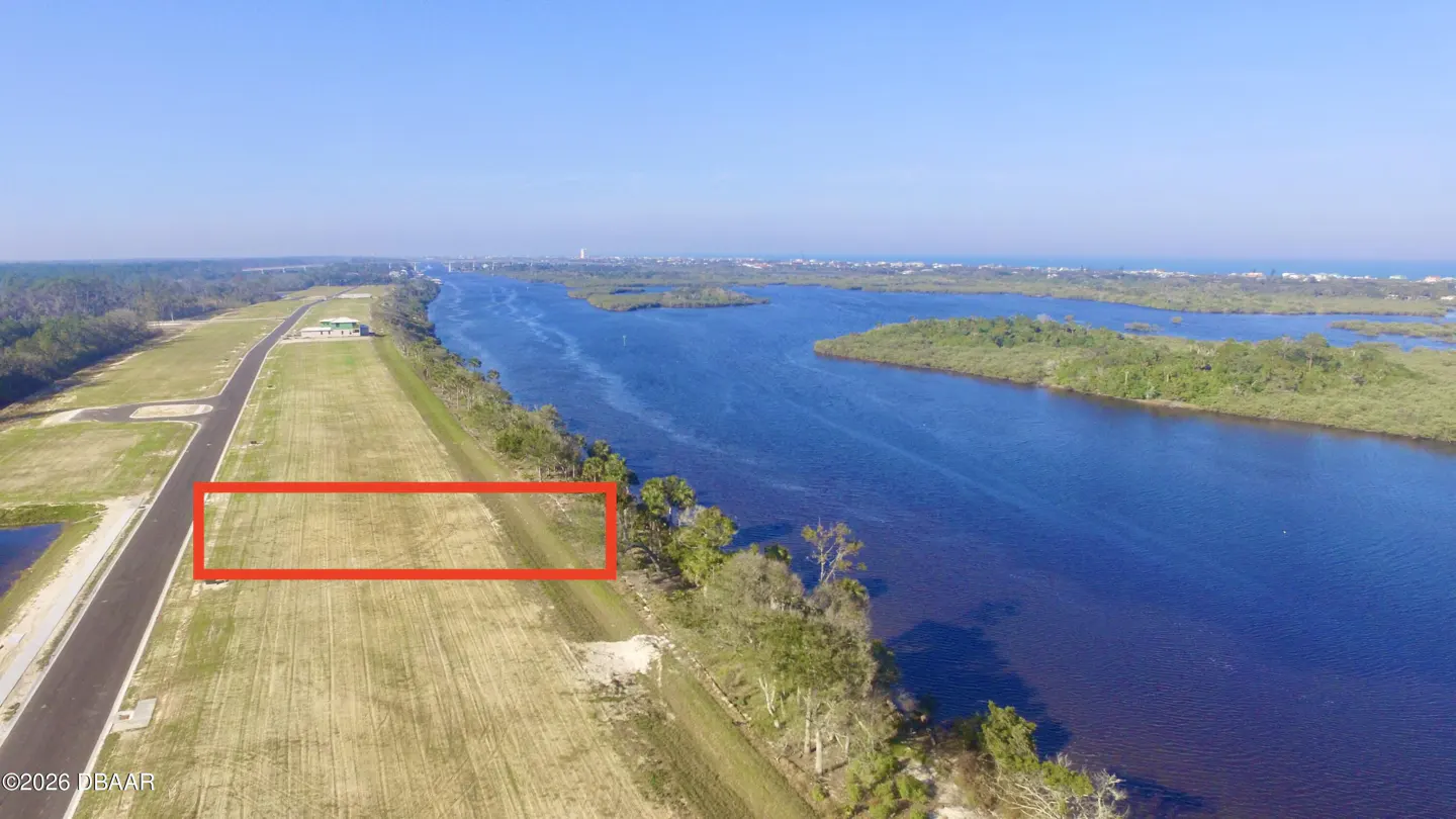 Aerial view of a grassy lot outlined in red, next to a blue river and green trees under a clear blue sky.