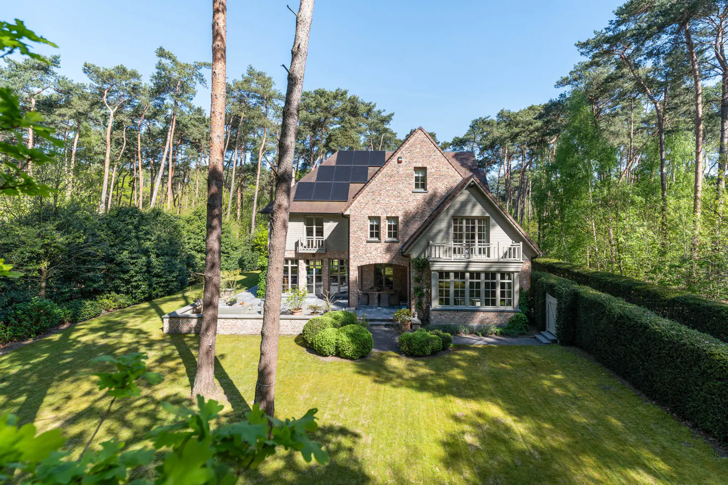 Two-story brick house with solar panels, surrounded by green lawn and trees under a blue sky.