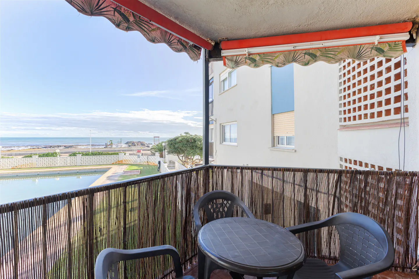 Balcony with a small table and two chairs, overlooking a swimming pool and the ocean under a red and patterned awning.