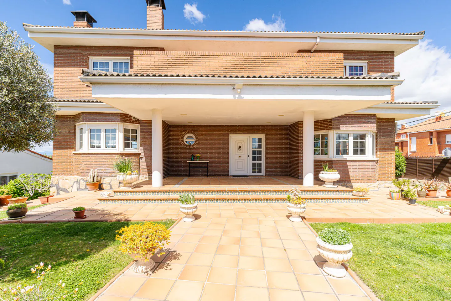Two-story brick house with a covered porch, white columns, and a tiled walkway lined with potted plants.