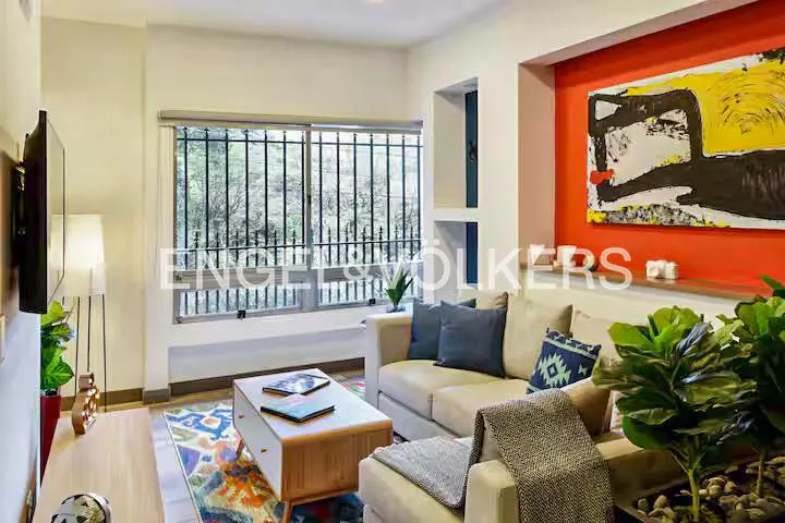 Living room with beige sofa, blue pillows, and a colorful rug. A large window with bars overlooks trees. An orange wall features abstract art.