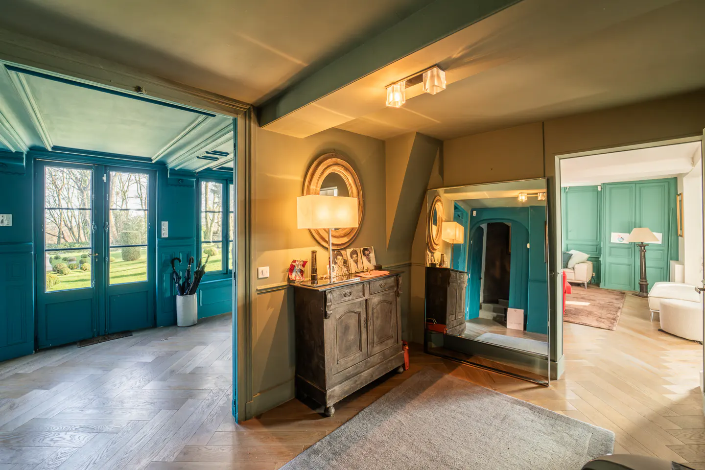 A foyer with a wooden floor, a gray rug, and a dark wood cabinet with a lamp and photos. A mirror reflects a teal room.