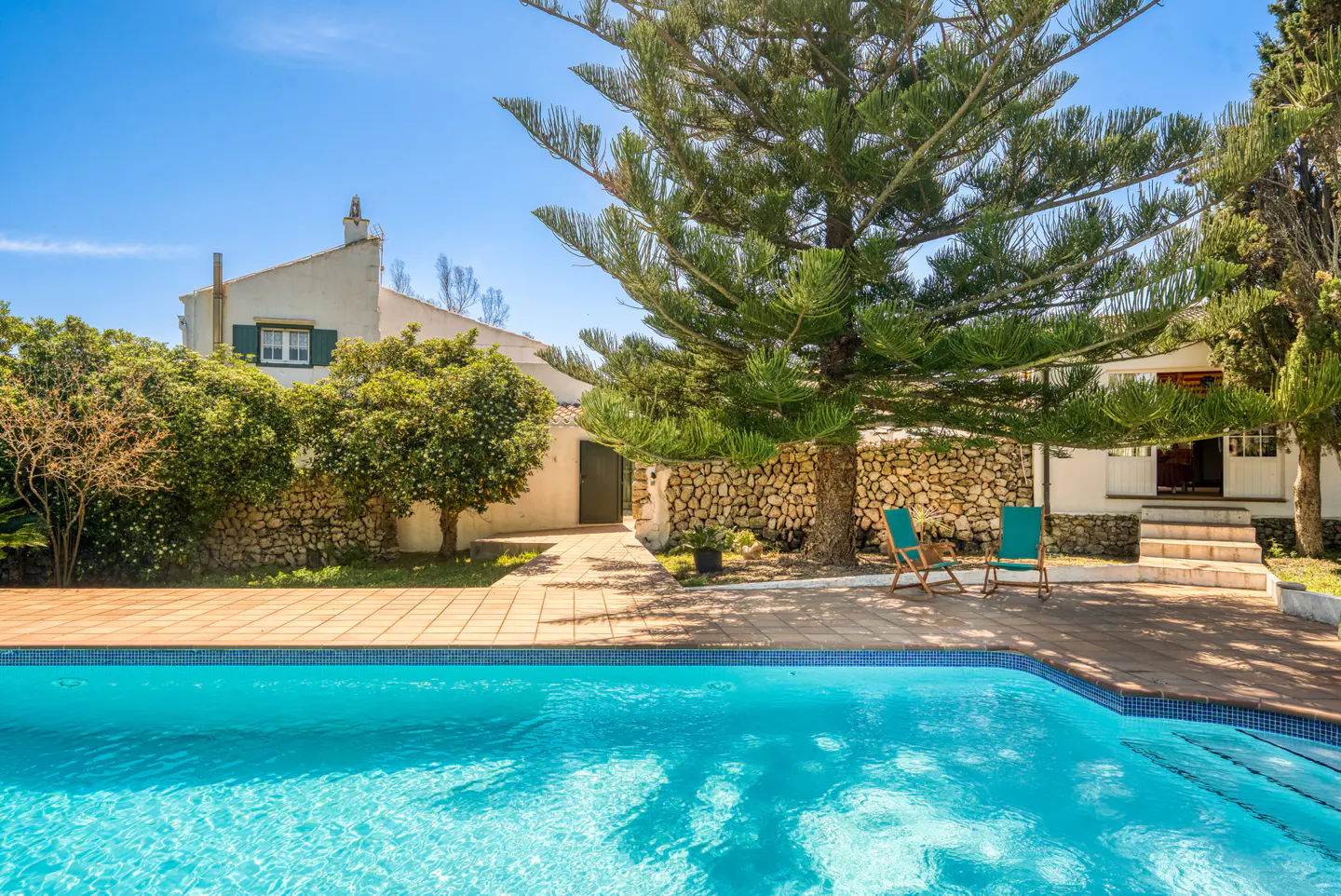 A bright blue pool in front of a white house with green shutters and a stone wall. Two teal chairs sit under a large pine tree.