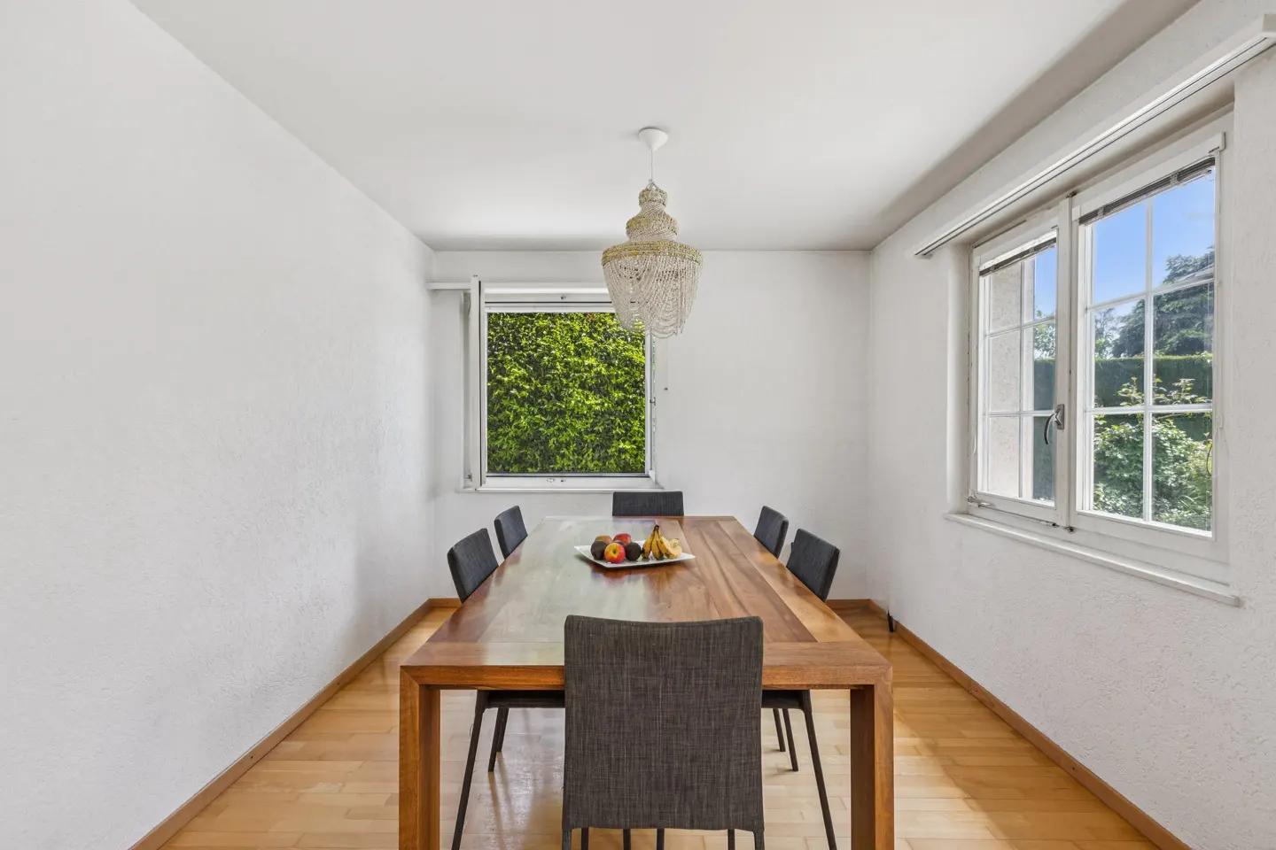 A dining room with a wooden table, six chairs, and a chandelier. Fruit sits on the table. Two windows show greenery outside.