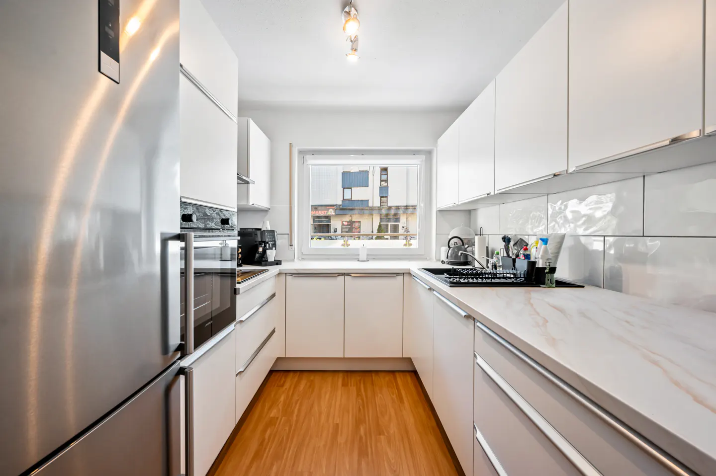 A modern kitchen with white cabinets, marble countertops, and stainless steel appliances. A window provides natural light.