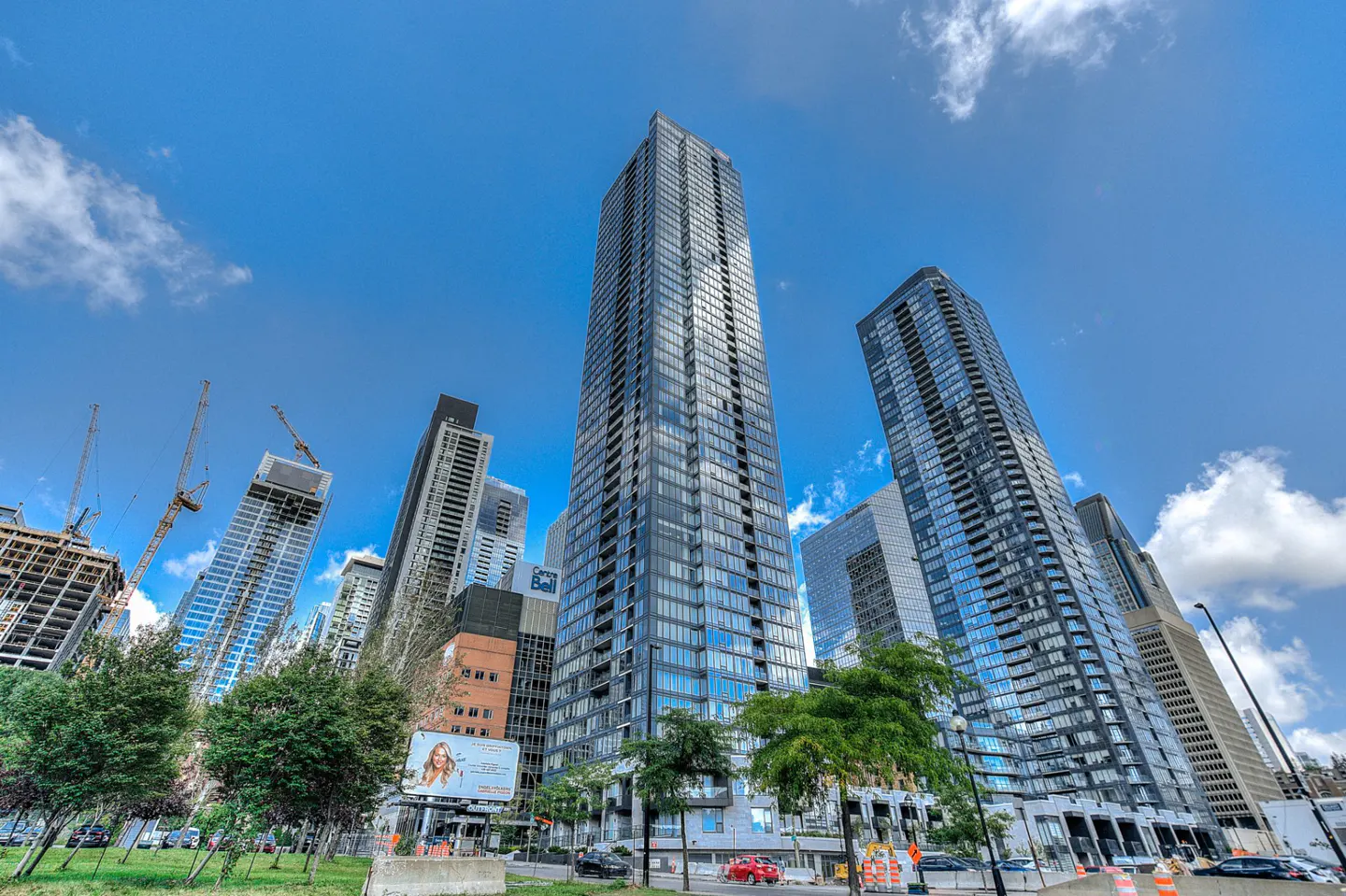Low-angle view of tall, modern glass skyscrapers against a blue sky with scattered clouds. Green trees line the street in front of the buildings.