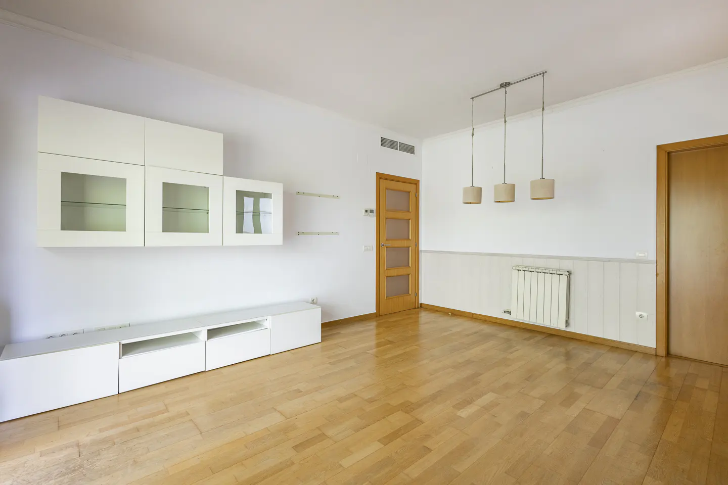 Bright, empty living room with wood floors, white walls, and built-in white cabinets. Three pendant lights hang from the ceiling.