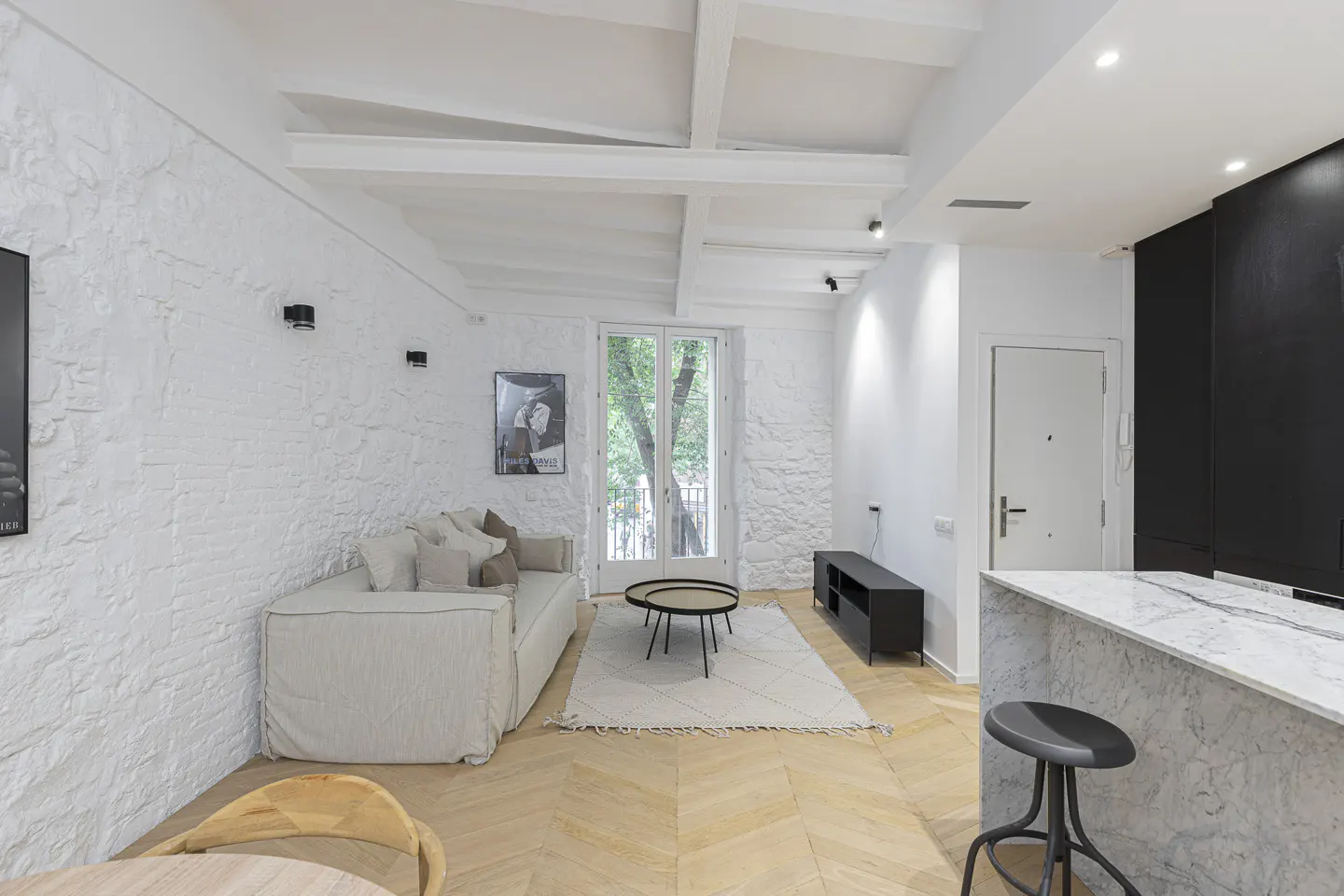 Bright living room with white textured walls, light sofa, and herringbone wood floors. Balcony doors let in natural light. Black kitchen accents.