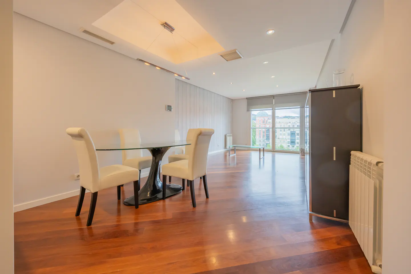 Bright dining room with hardwood floors, a glass table with four cream chairs, and a large window overlooking a city.