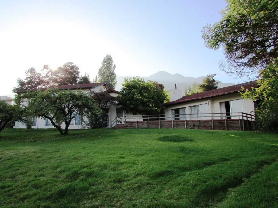 A white house with a red roof sits on a green lawn with trees and mountains in the background.