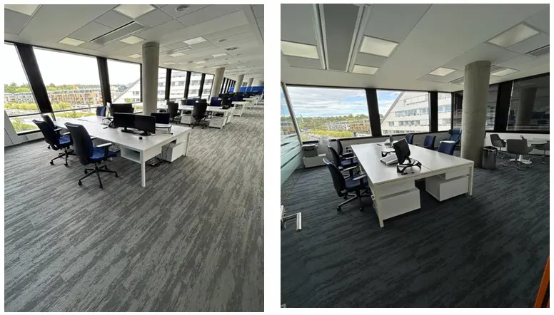 Open office space with white desks, blue chairs, and gray carpet. Large windows offer a city view.