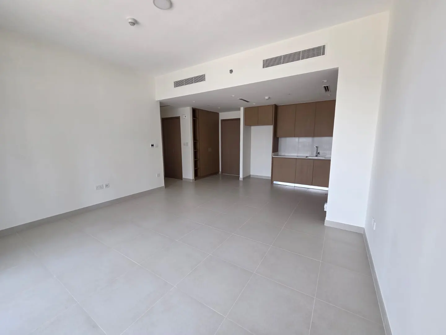 An empty, bright apartment with white walls, gray tile floors, and a brown kitchen in the background.