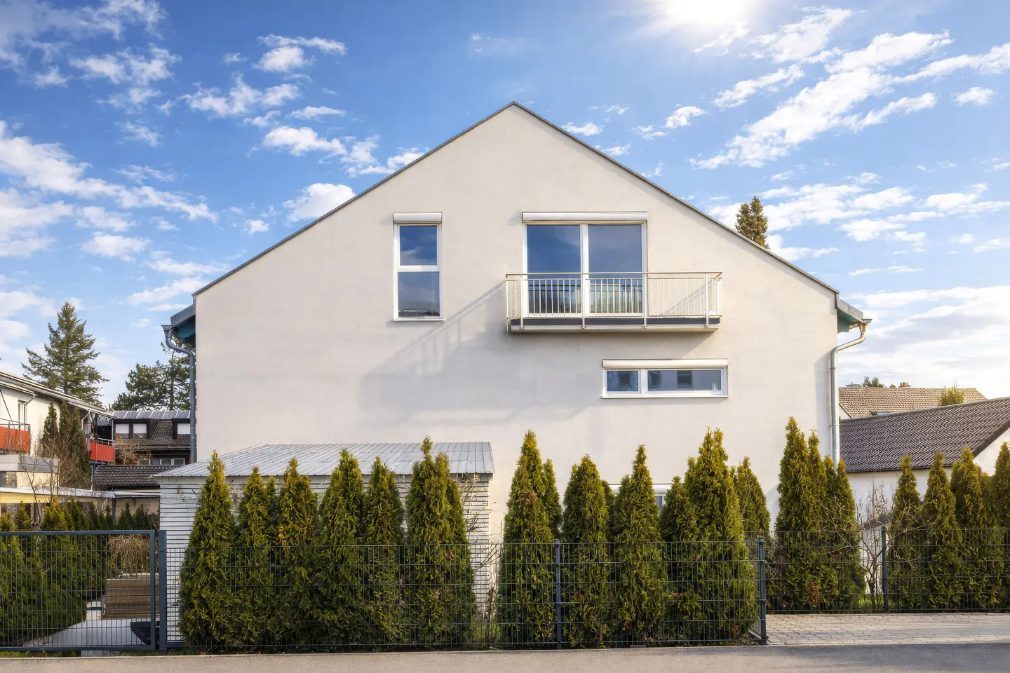 Modern two-story house with a balcony, white facade, and a row of green trees in front of a black metal fence. Blue sky with clouds.