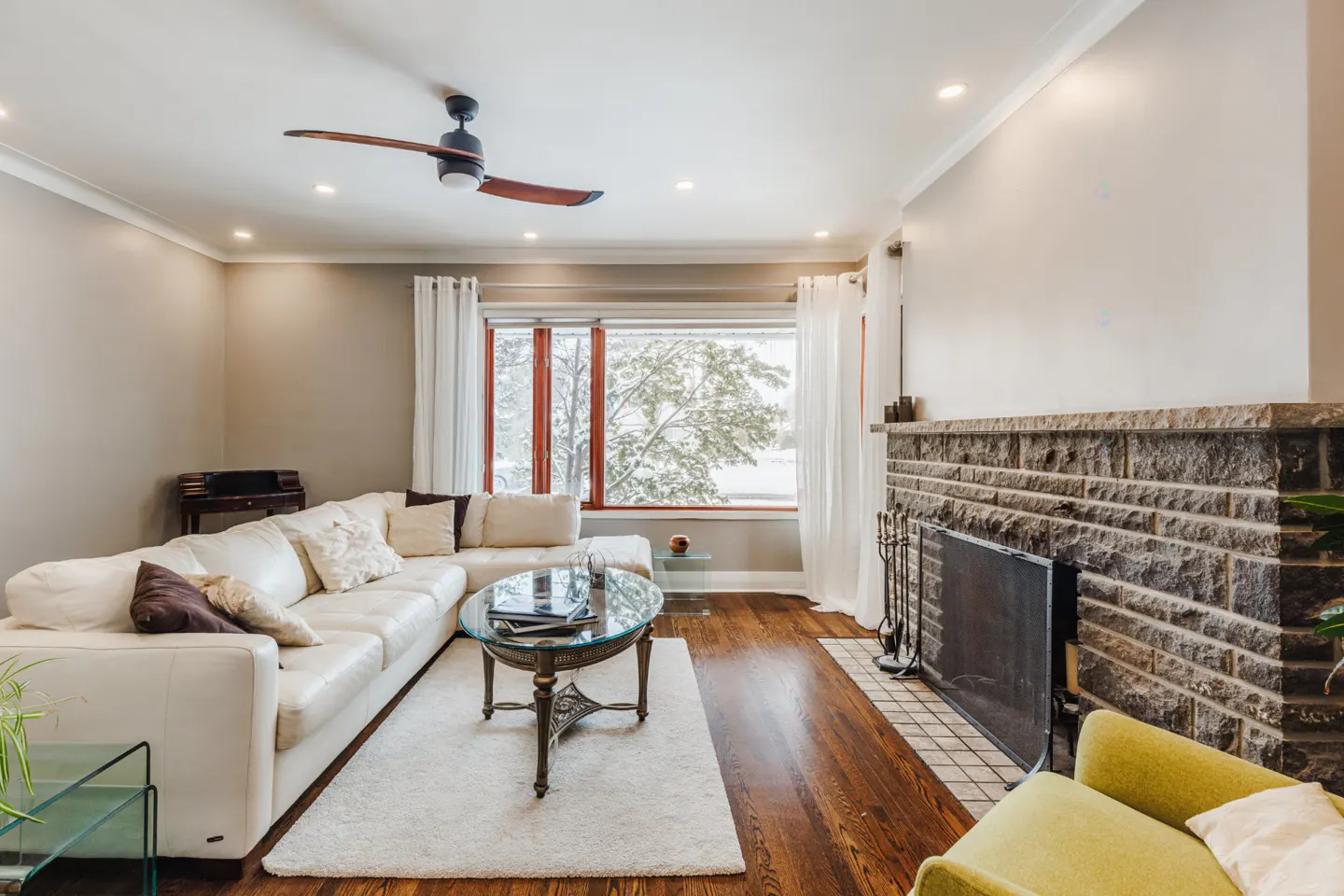 Living room with a white sectional sofa, glass coffee table, stone fireplace, and a window view of a snowy landscape.