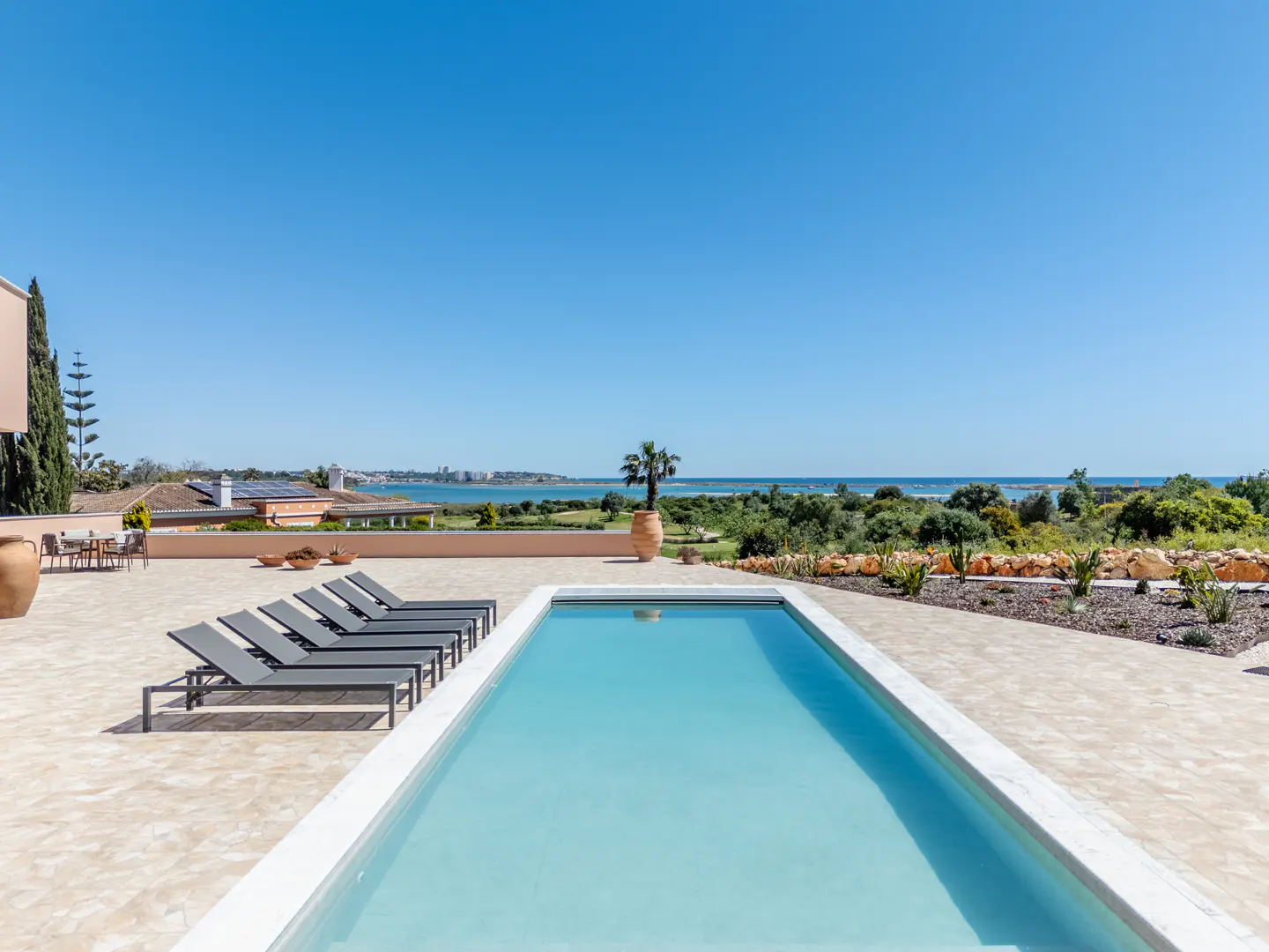 A rectangular pool with lounge chairs on a patio overlooking the ocean under a clear blue sky.