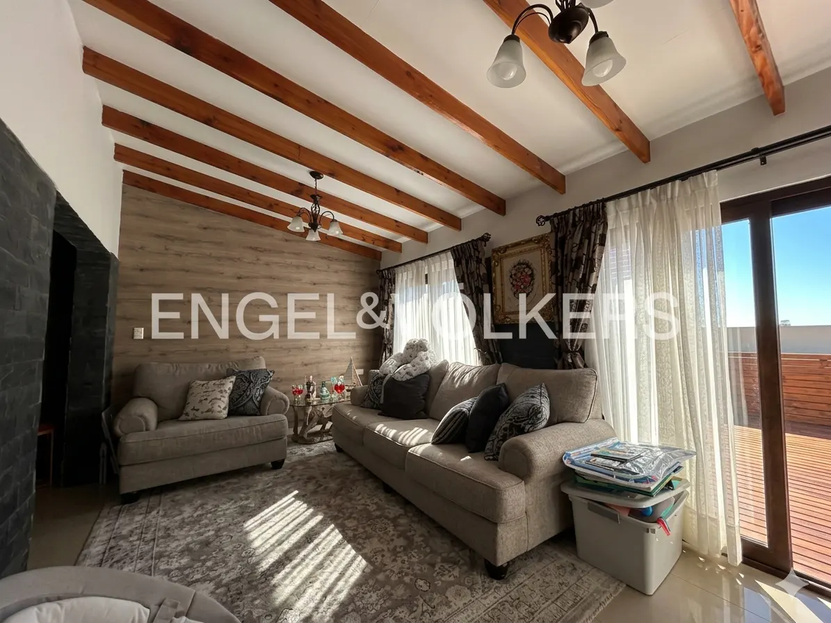 Living room with beige sofa, loveseat, and patterned rug. Wooden beams accent the ceiling. Engel & Volkers logo is visible on the wall.