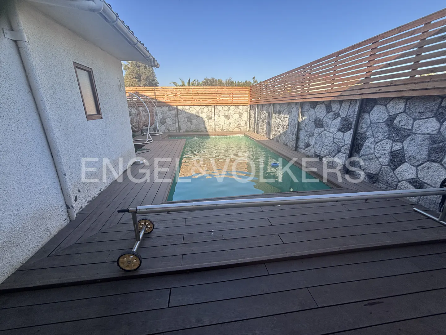 Outdoor pool with wooden deck and stone wall. Pool cover on rollers is partially visible. Blue sky.