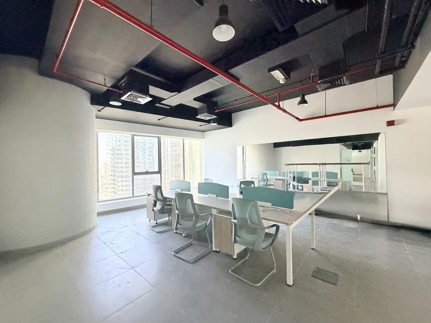 Bright, modern office space with desks, chairs, and large windows. Black ceiling with red pipes and city view.