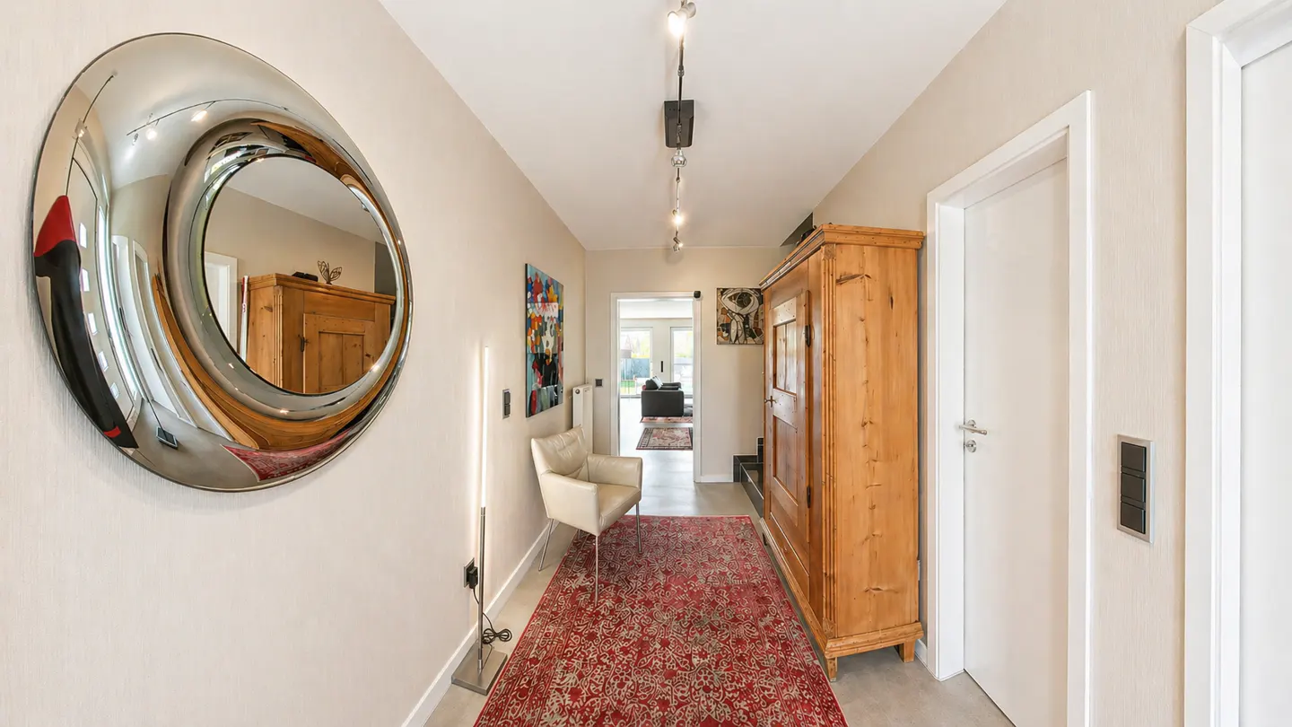 Hallway with a large round mirror, red rug, wooden cabinet, and a white chair. A door leads to another room.