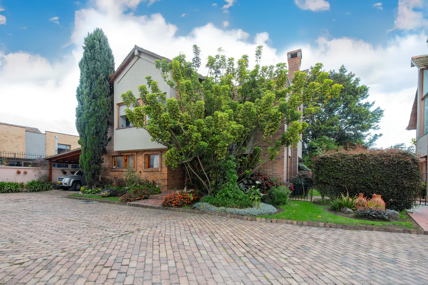 Exterior view of a two-story brick house with a gray second story, surrounded by lush greenery and a brick driveway.