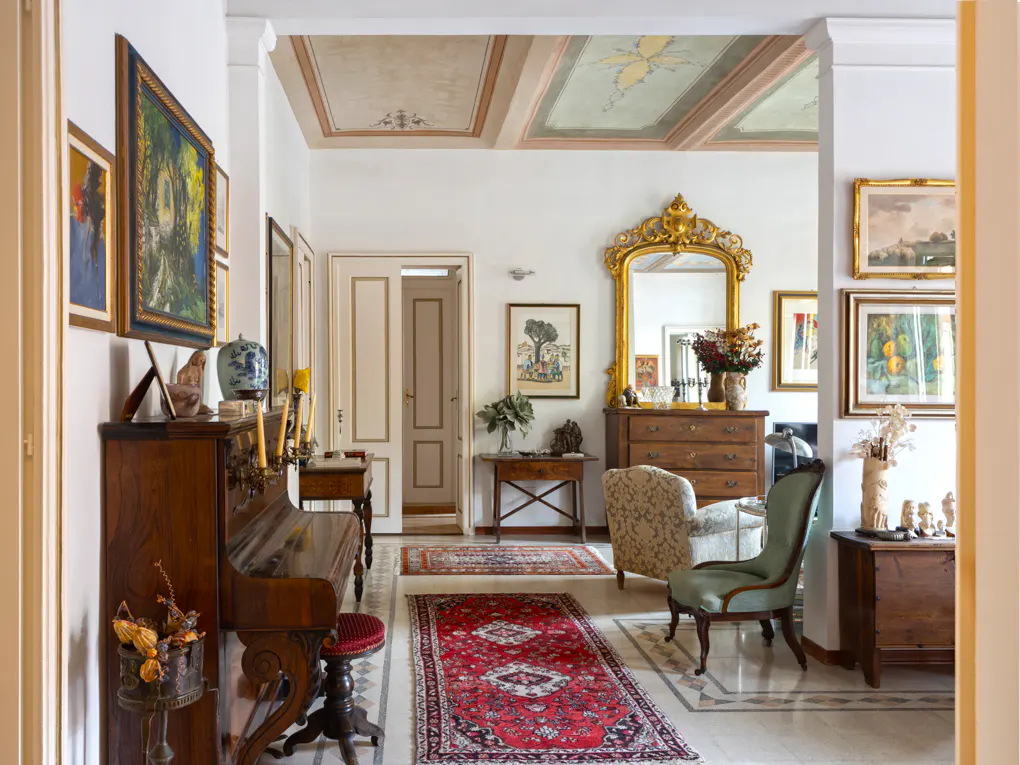 Elegant hallway with marble floors, red rugs, and antique furniture. Walls are adorned with paintings, and the ceiling has decorative panels.