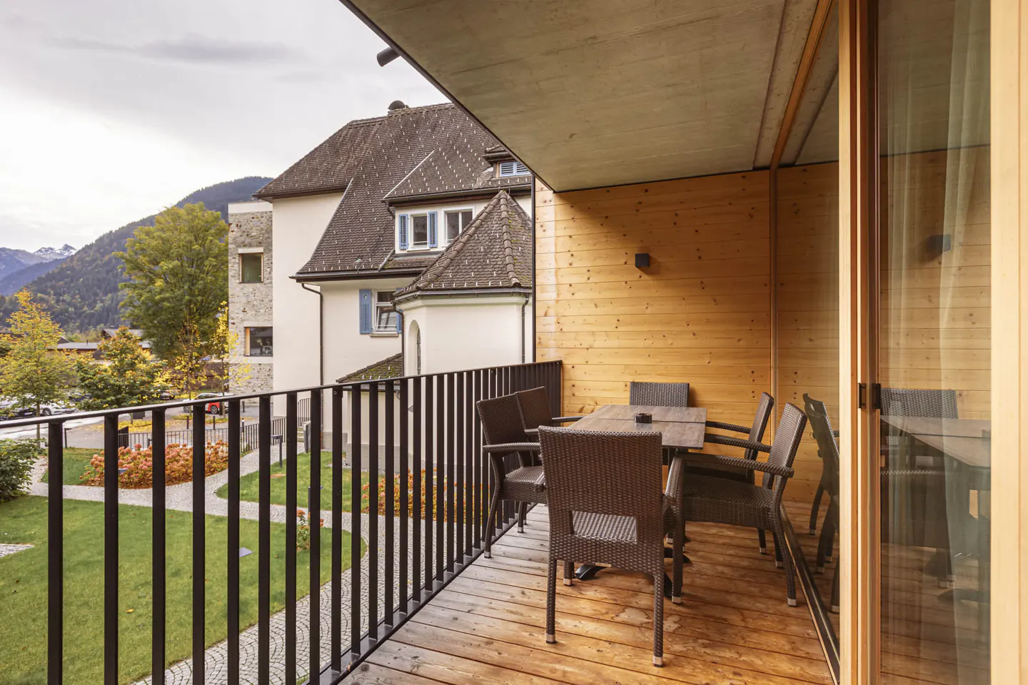 Modern balcony with wooden walls and floor, featuring a dining set overlooking a garden and traditional mountain buildings.