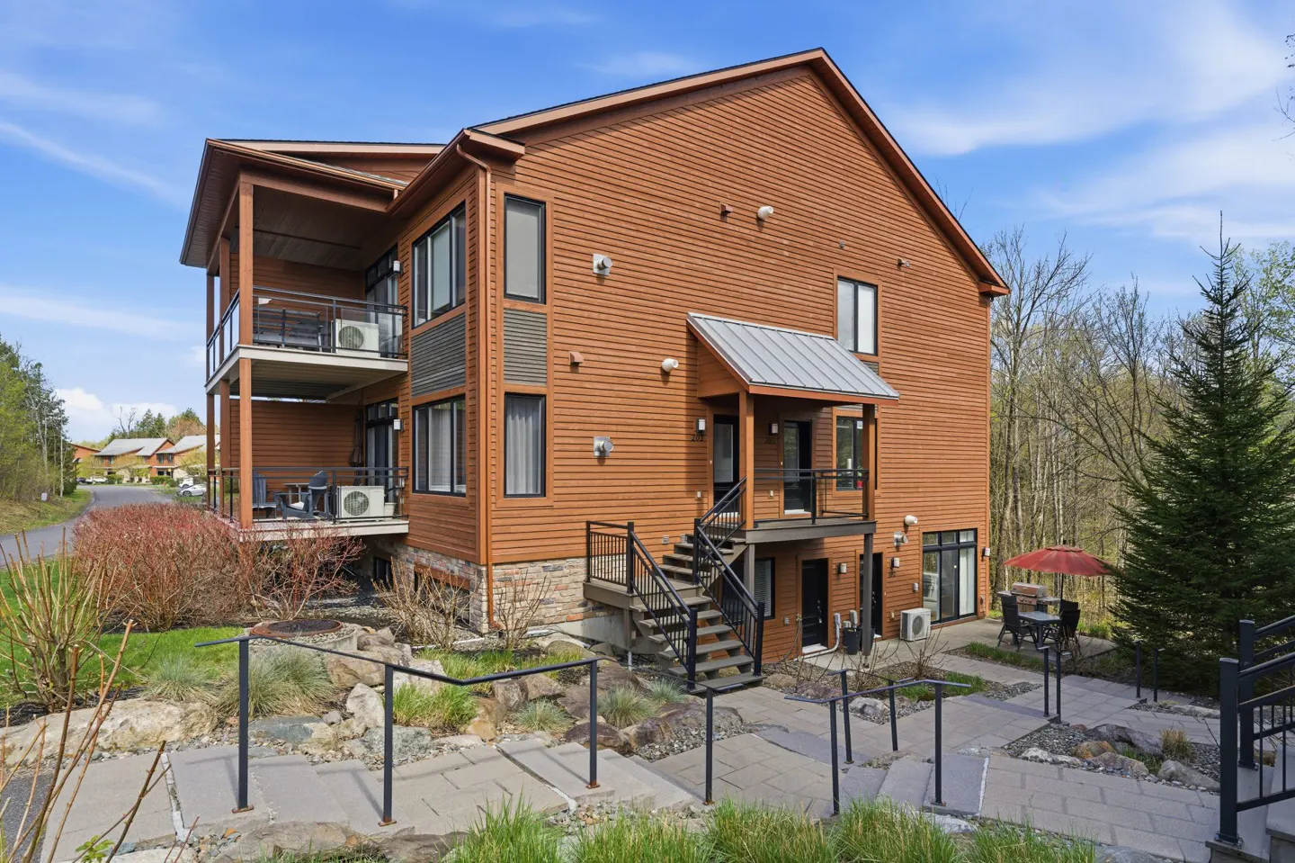 Exterior view of a three-story brown wood-sided building with black framed windows and balconies, stone walkway and landscaping.