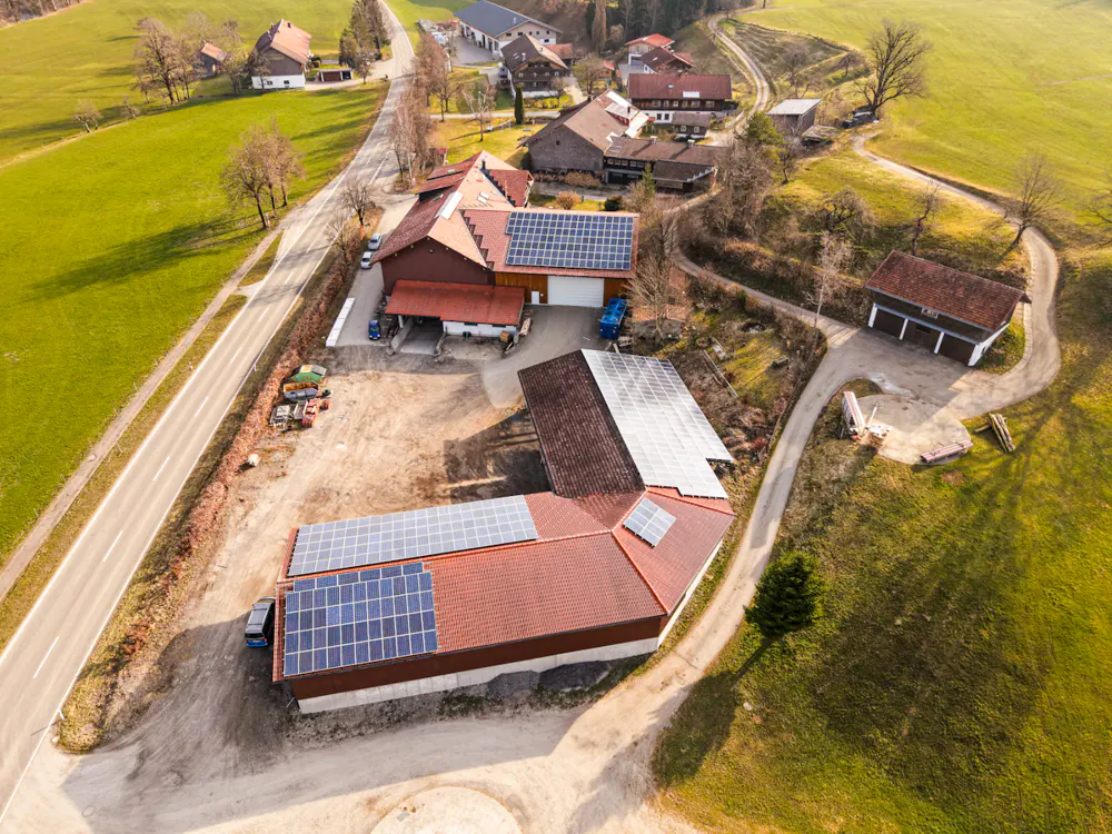 Aerial view of a farm with solar panels on red roofs, surrounded by green fields and a road.