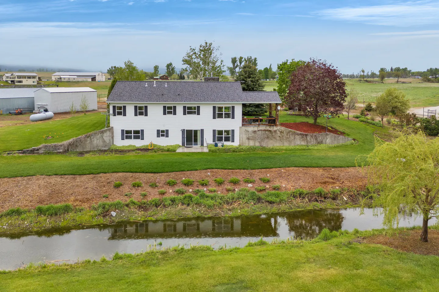 Two-story white house with black shutters, gray roof, and covered patio. A stream runs in front of the house, with green grass and trees surrounding it.