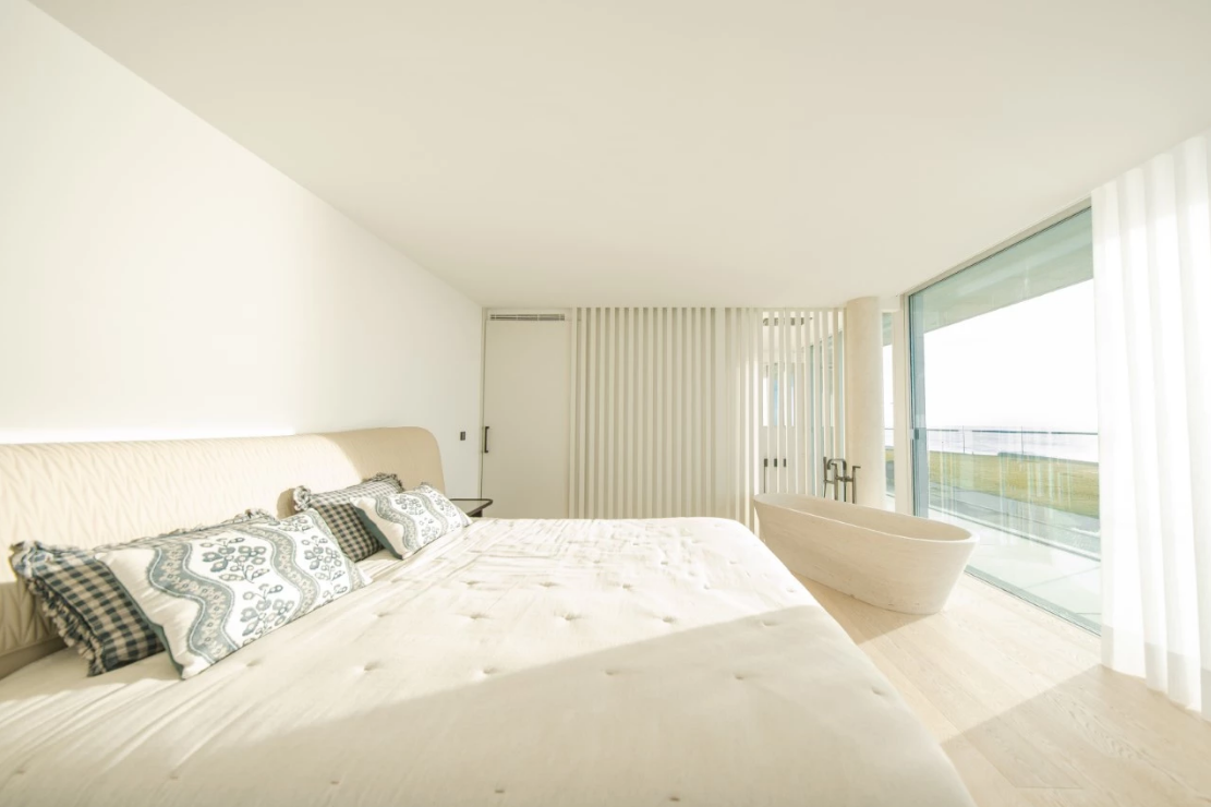 Bright, modern bedroom with a large bed, patterned pillows, and a freestanding tub near a window with a view.