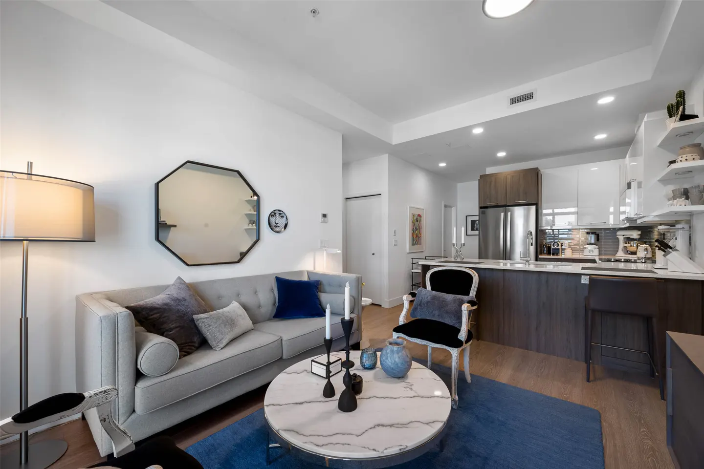 Bright apartment living room with a gray sofa, marble table, blue rug, and open kitchen with stainless steel appliances.