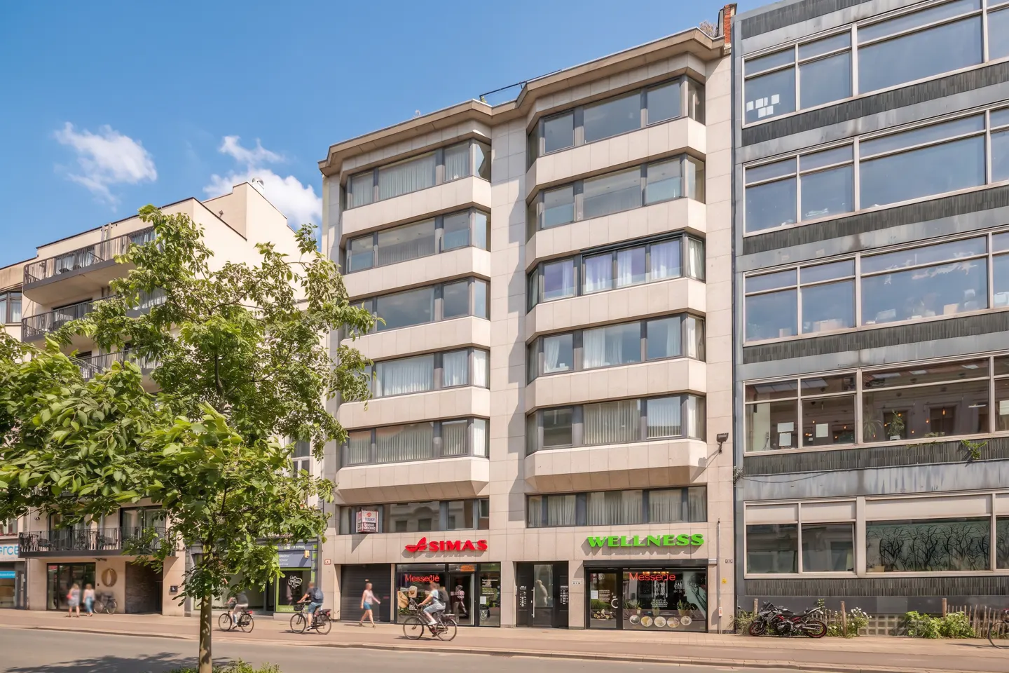 Street view of a multi-story building with shops on the ground floor. People are walking and cycling on the sidewalk.