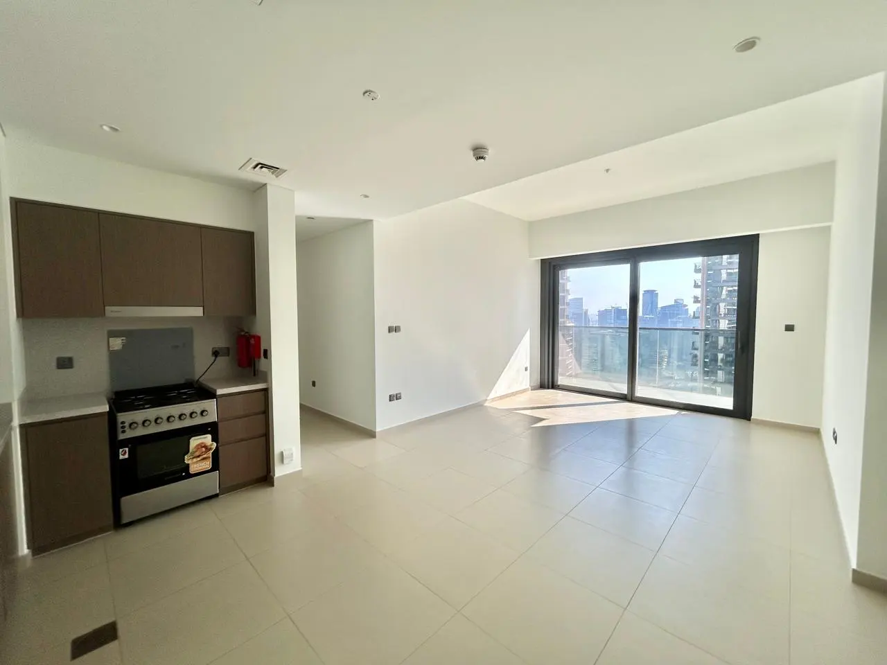 Bright, empty apartment with beige tile floors, white walls, and brown cabinets. A large sliding glass door shows a city view.