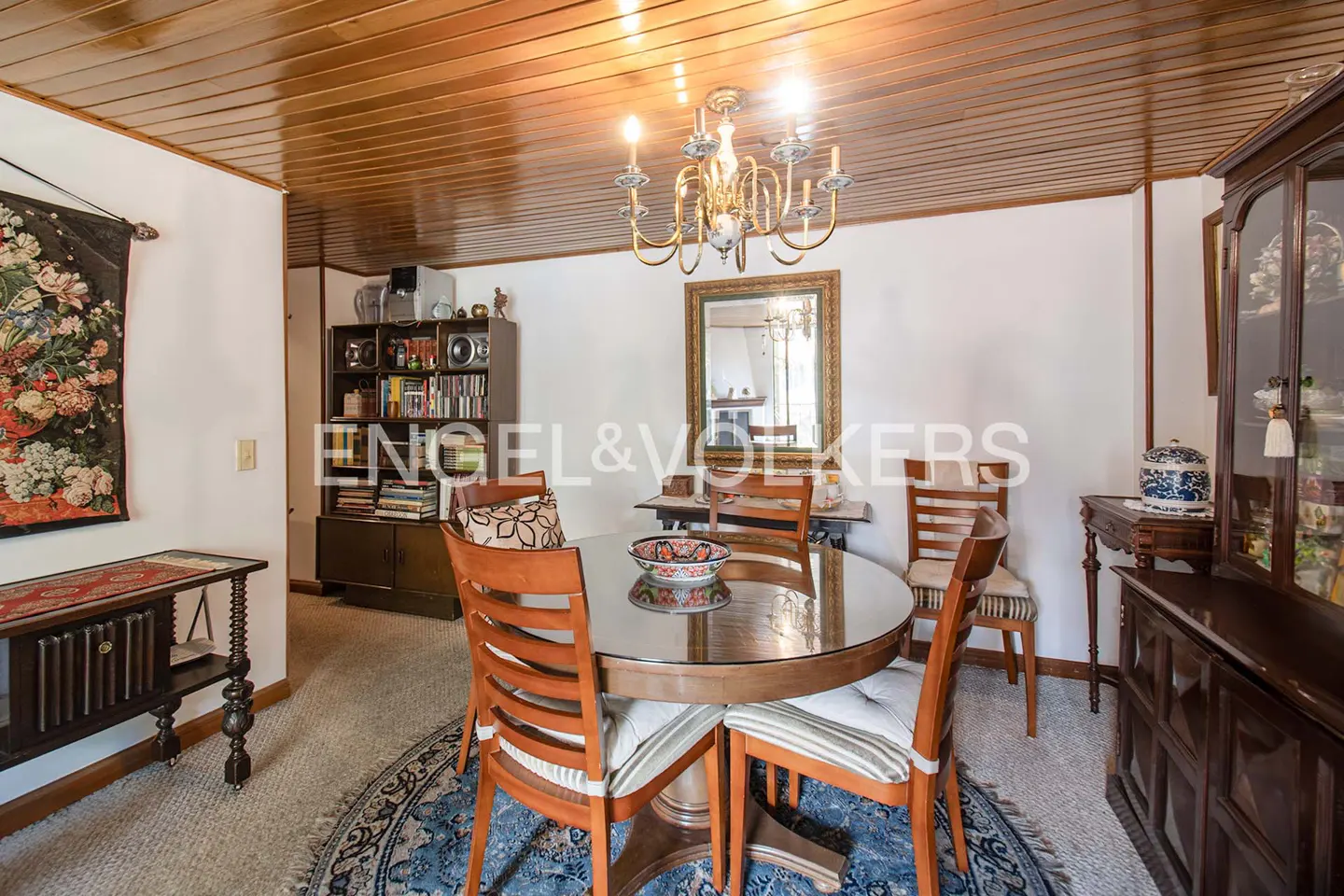 A dining room with a round table, wooden chairs, and a chandelier. A bookshelf and a china cabinet are visible.