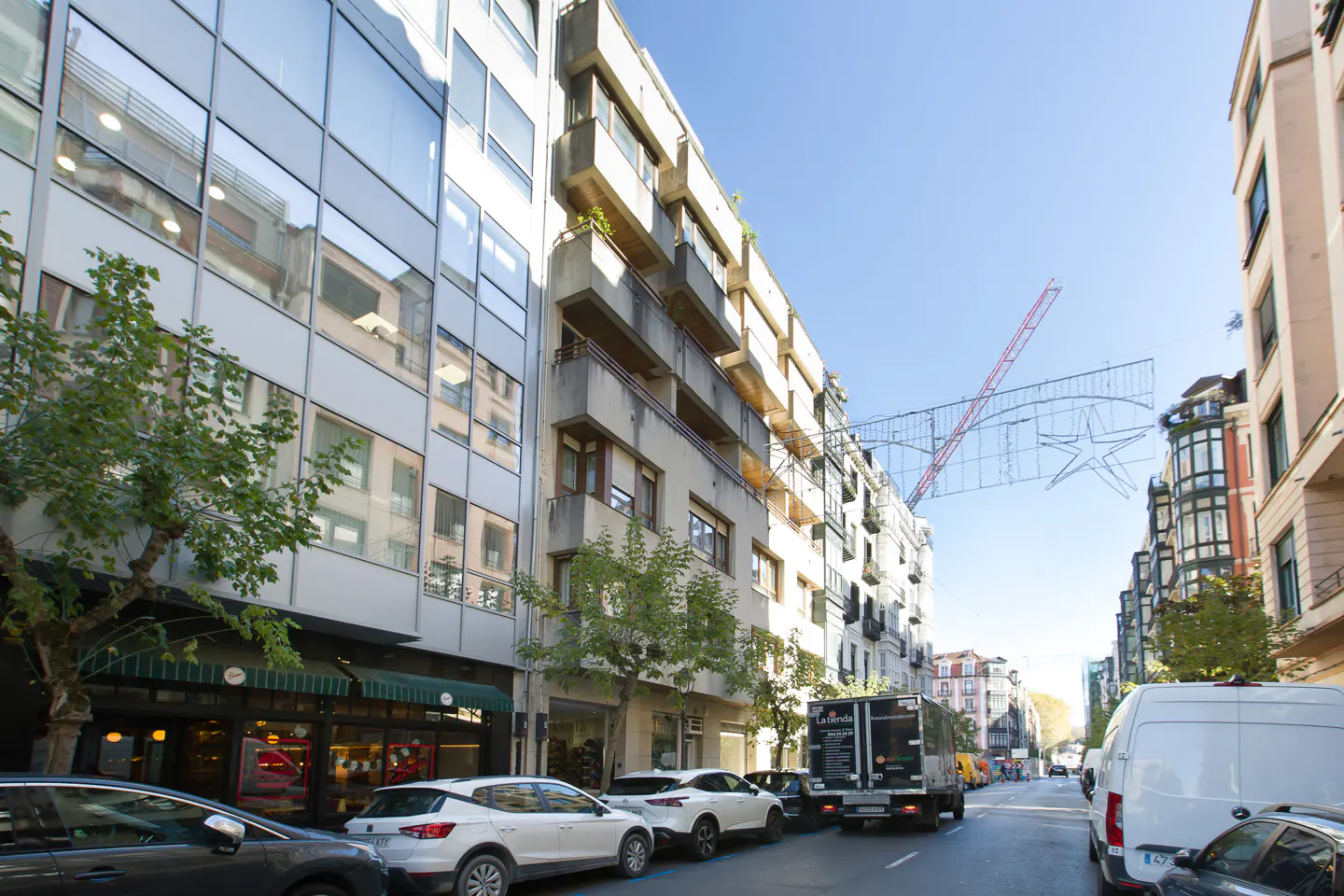 Street view of buildings with cars parked along the side of the road. Christmas decorations hang above the street.