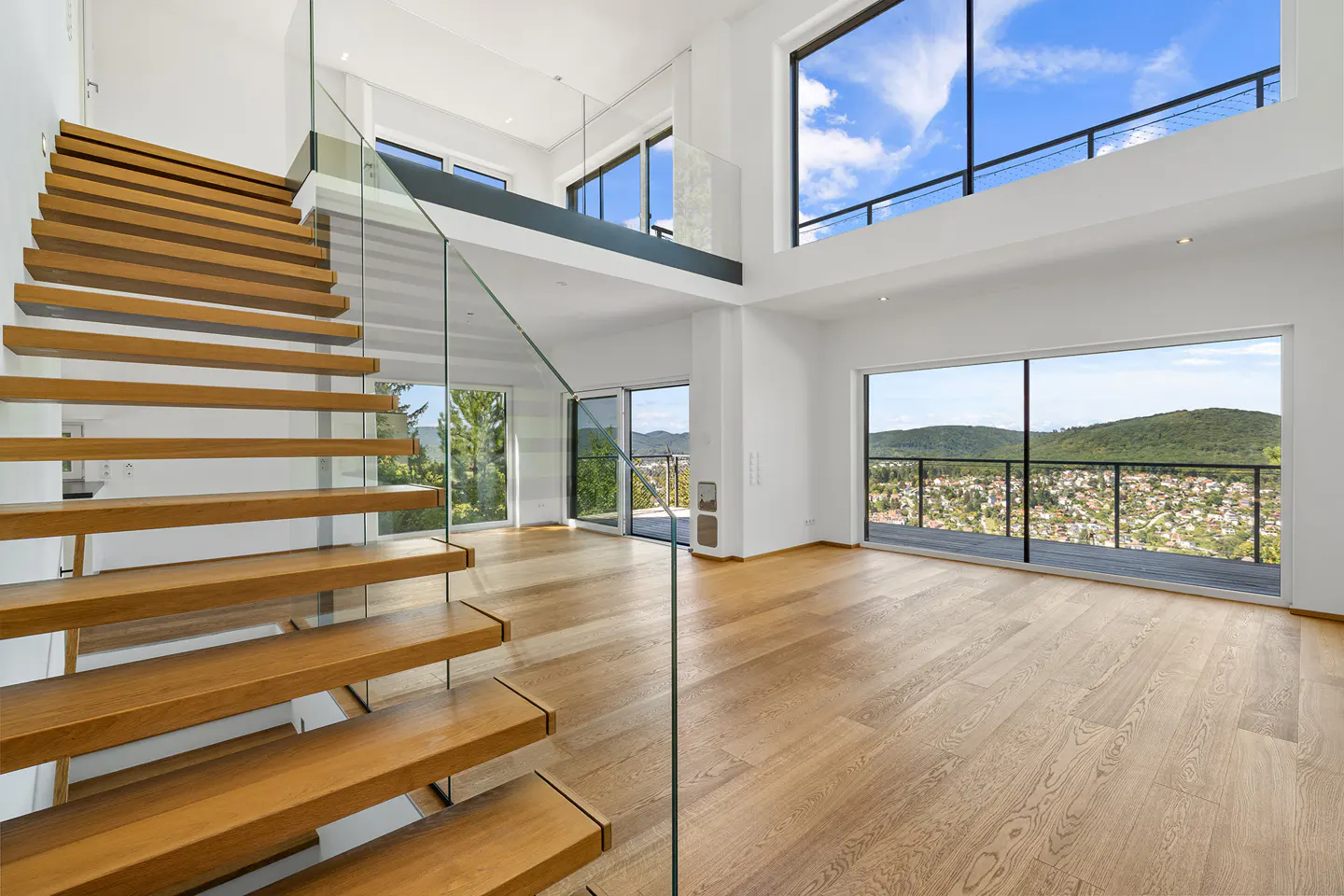 Bright, modern living room with wood floors, open staircase with glass railing, and large windows overlooking a green landscape.