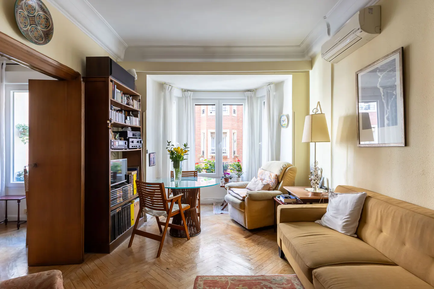 Cozy living room with a yellow sofa, armchair, glass table, and bookshelf. Natural light streams through the bay window.
