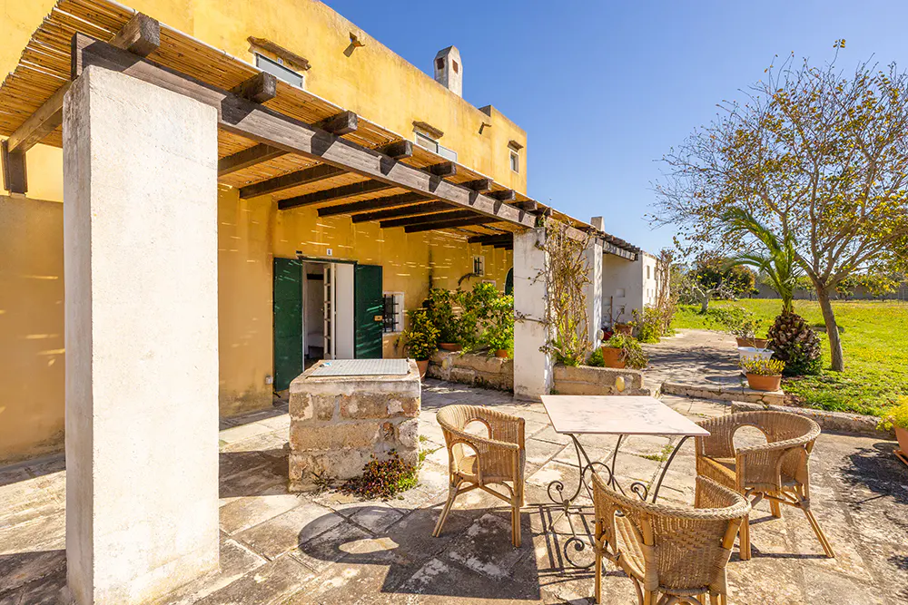Outdoor patio with wicker chairs and a metal table on stone tiles. A yellow building with green shutters is in the background.
