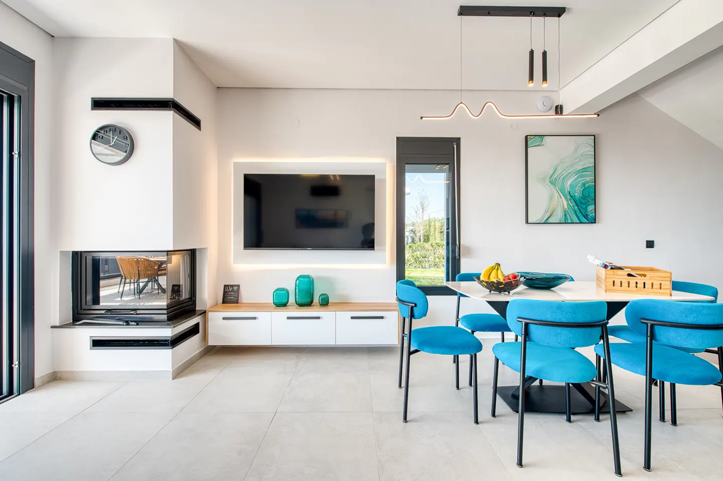 Bright, modern living room with a fireplace, TV, and dining table with blue chairs. A painting hangs on the wall.