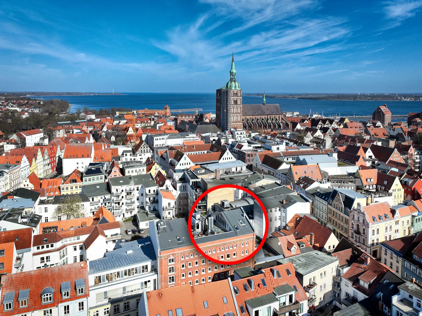 Aerial view of a European city with red-tiled roofs, a church steeple, and a red circle highlighting a building.