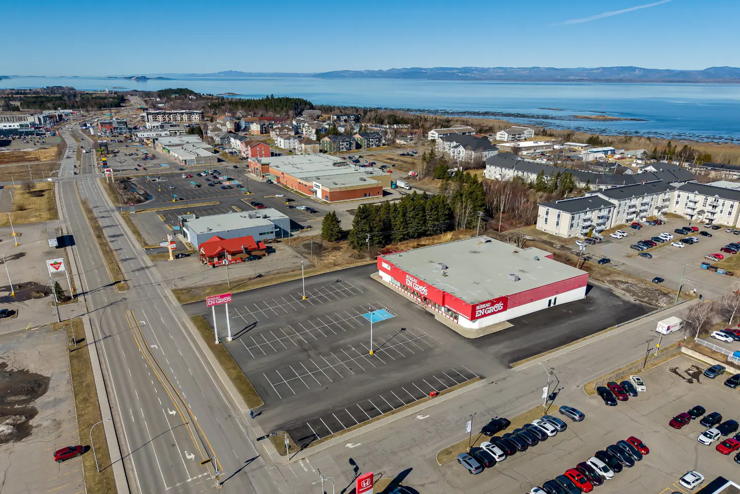 Aerial view of a commercial area with stores, parking lots, and a road, with a lake and mountains in the background under a blue sky.