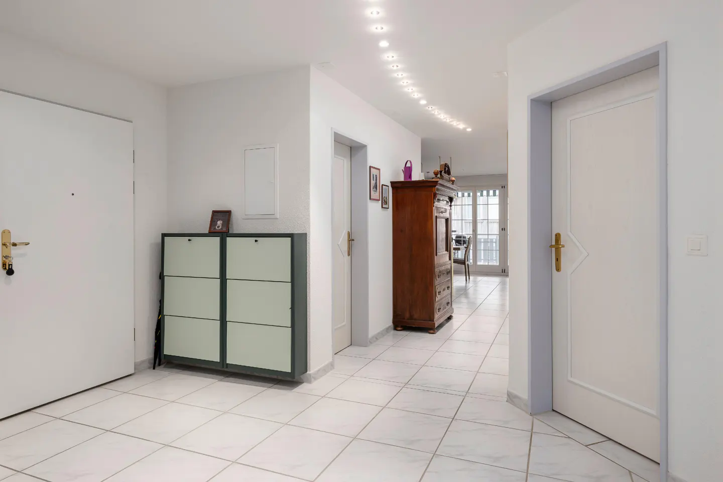 Bright hallway with white walls, tiled floor, and recessed lighting. A shoe cabinet and antique wooden dresser furnish the space.