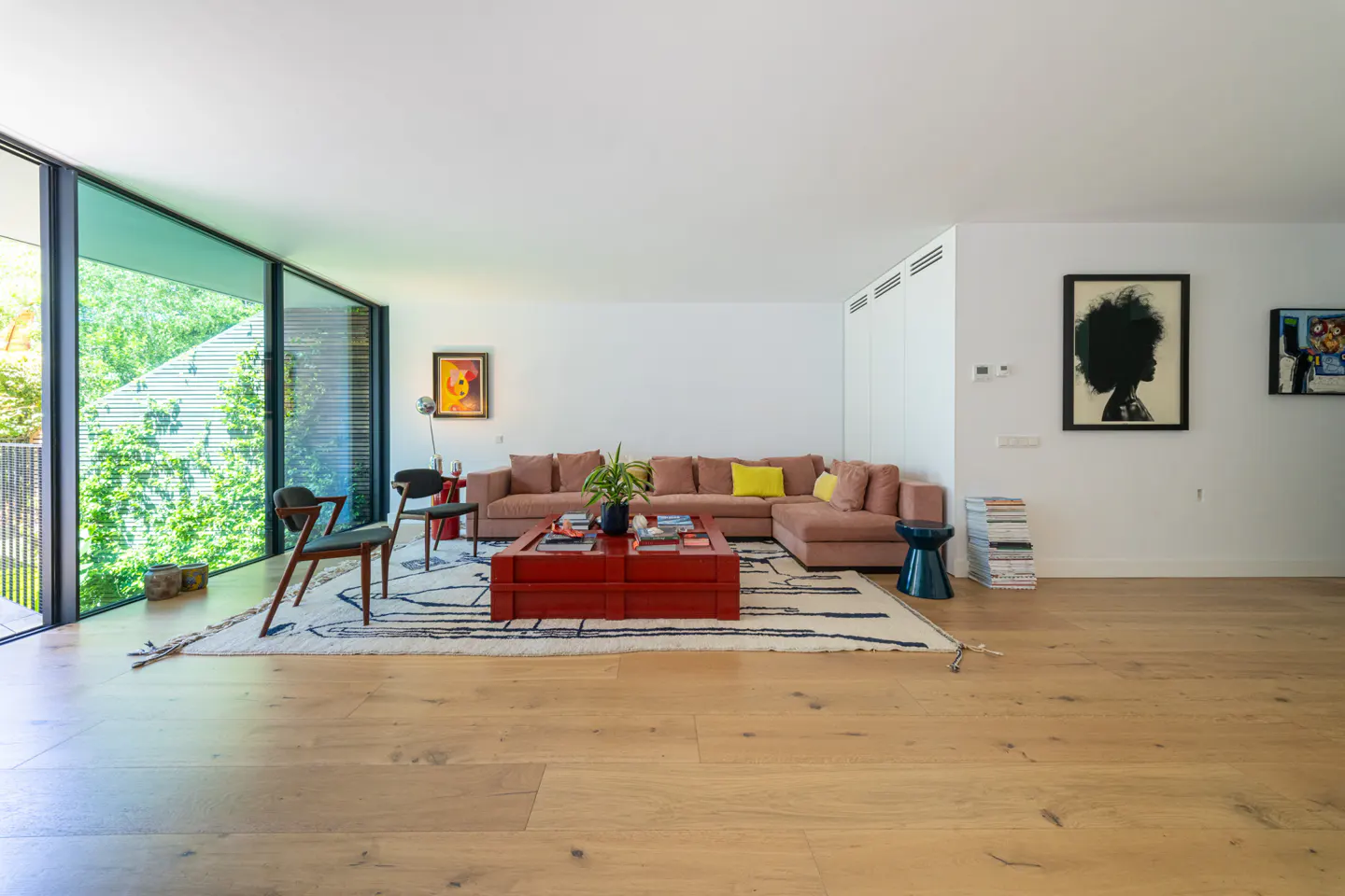 Modern living room with a pink sectional sofa, red coffee table, and large windows overlooking lush greenery.