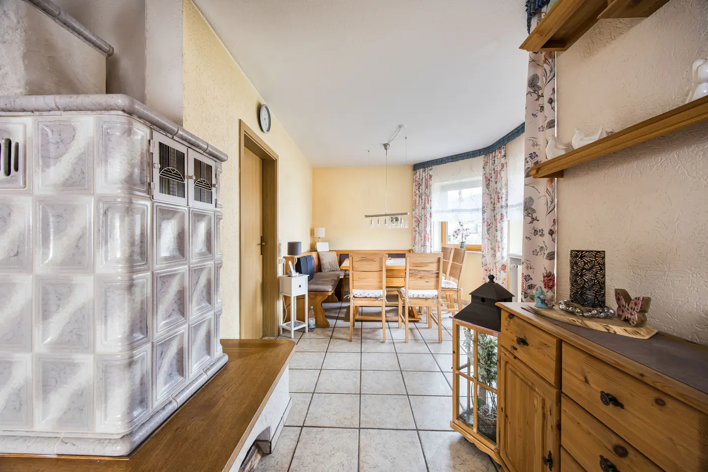 Living room with a white tiled stove, wooden dining table and chairs, and a wooden dresser with shelves.