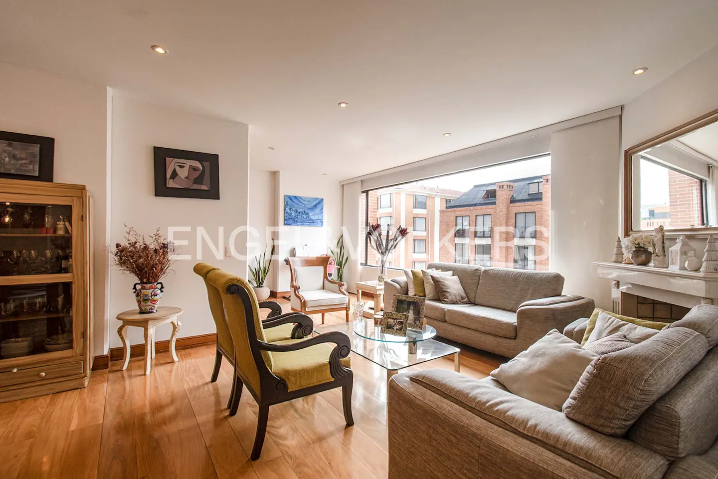 Bright living room with hardwood floors, beige sofa, two yellow armchairs, glass coffee table, and large city view window.