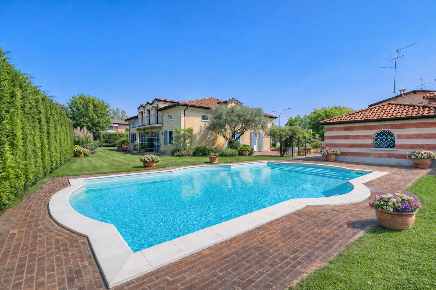 Outdoor pool with clear blue water, surrounded by brick patio, lawn, and a yellow house under a clear blue sky.