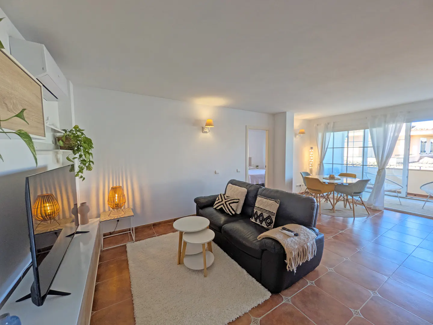 Bright living room with a black sofa, white rug, and dining area visible through large windows. Brown tile flooring and white walls.