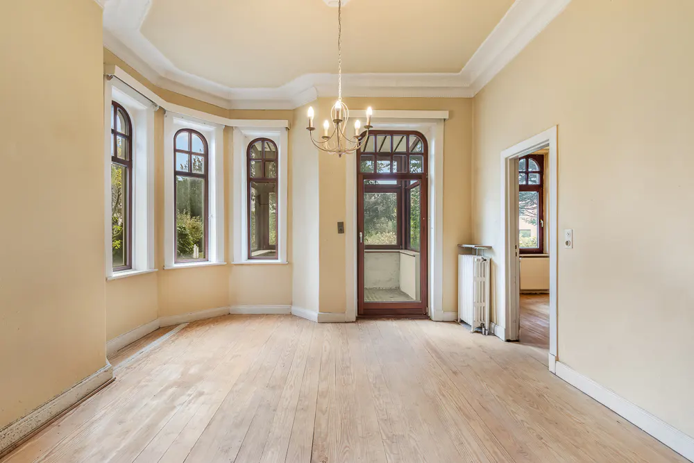 Empty room with light yellow walls, wood floors, and a chandelier. Arched windows and a door lead to the outside.