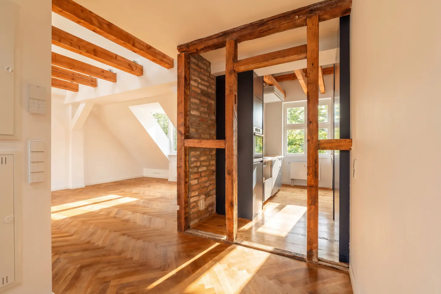Interior view of an apartment with herringbone wood floors, exposed beams, and a kitchen visible through a framed opening.